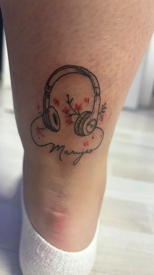 Tatuaje fine line de unos auriculares con flores y un nombre en la pierna
